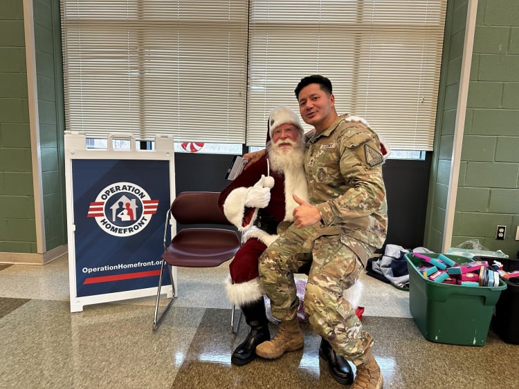 A person in military camouflage uniform sitting on Santa Claus's lap next to a sign that reads "Operation Homefront" with the website OperationHomefront.org, in a room with green walls and window blinds.