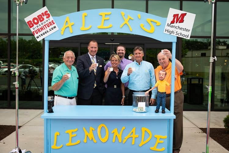 Alex's Lemonade stand with six people holding cups behind a blue lemonade booth. Signs read "BOB'S Discount Furniture MYBOBS.COM" and "Manchester High School." A clear jar with money is on the counter.