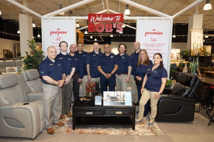 Group of employees wearing navy blue polo shirts standing in a furniture store under a sign reading "Welcome to Bob's" with banners stating "Our purpose" and "Our promise."