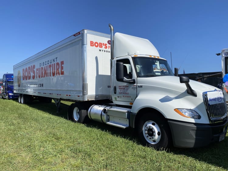 White semi-truck with trailer branded "Bob's Discount Furniture" in red and black text, parked on grass under clear blue sky.
