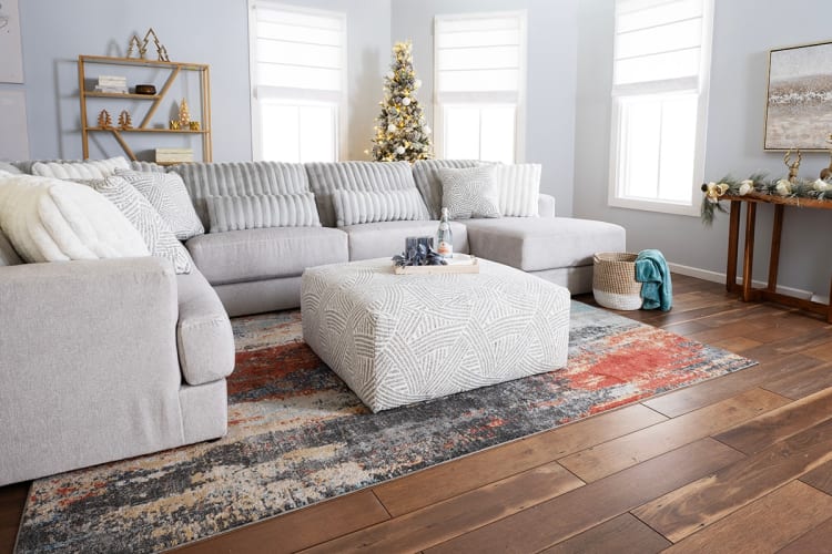 Modern living room with light gray sectional sofa, textured beige ottoman, multicolor abstract rug, wood flooring, and a decorated Christmas tree.