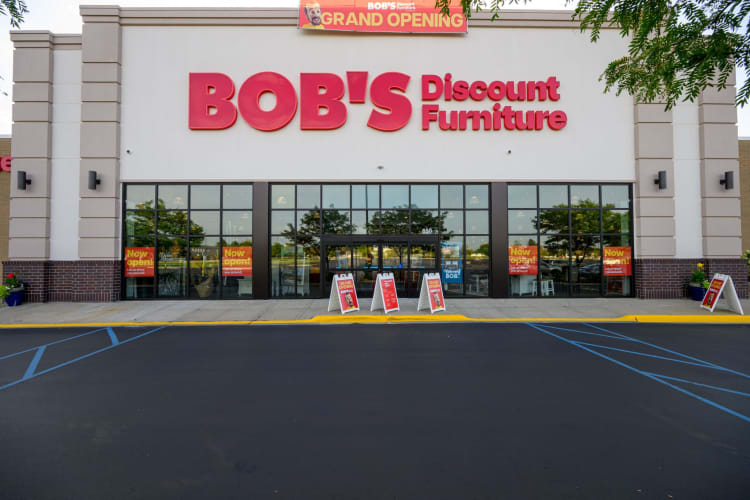 BOB'S Discount Furniture storefront with large red signage, glass entrance, and multiple "Now open!" and "Grand Opening" promotional signs.