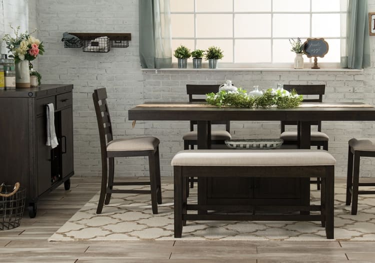 Dark wood dining set with beige cushioned bench and chairs on patterned rug, featuring greenery centerpiece and bright window backdrop.