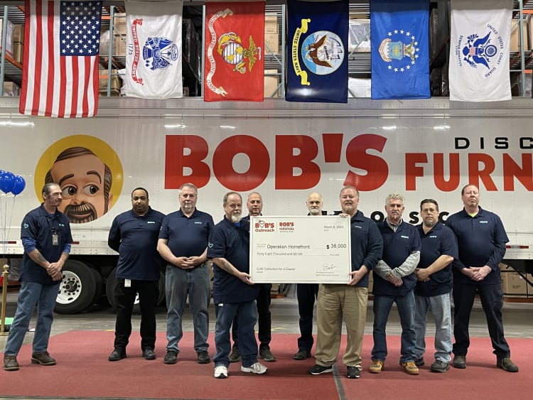Group of people standing in front of a Bob's Discount Furniture truck holding a large check for $38,000 made out to Operation Homefront, dated March 8, 2023. Six flags hang above the truck, including the American flag and military branch flags. The scene highlights a charitable donation event supporting veterans.