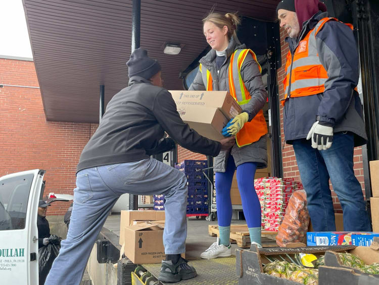 Workers wearing orange safety vests and gloves unload cardboard boxes labeled "KEEP REFRIGERATED" from a delivery truck at a loading dock with pallets of produce nearby.