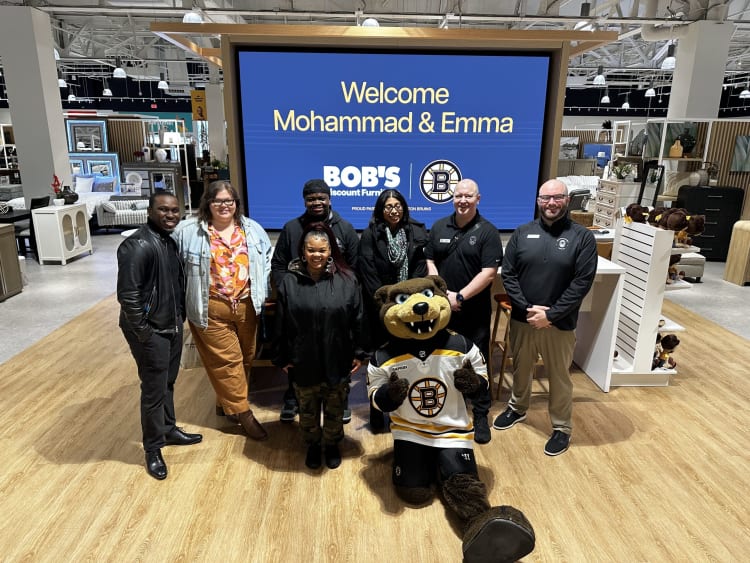 Welcome sign for Mohammad & Emma at Bob's Discount Furniture with a group of seven people and a Boston Bruins mascot in a store setting.