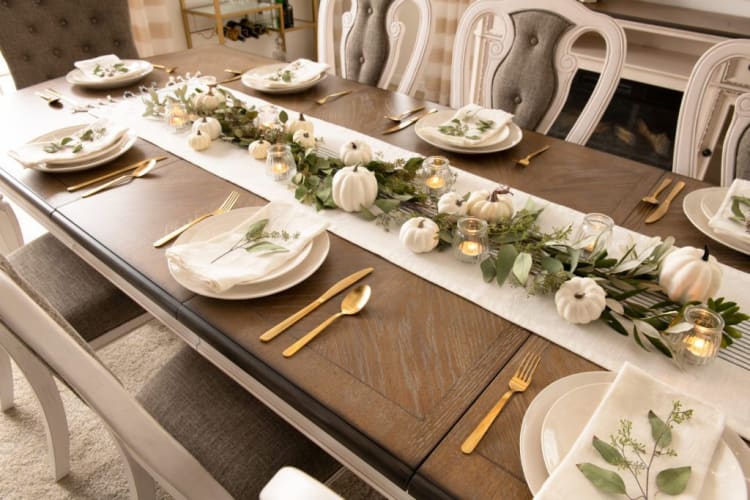 Dining table set with beige upholstered chairs, decorated with white mini pumpkins, green foliage, and lit candles on a white table runner.