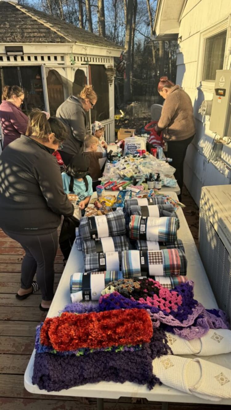 A long table outdoors displaying folded plaid blankets, colorful crocheted scarves, and white slippers, with people browsing items.