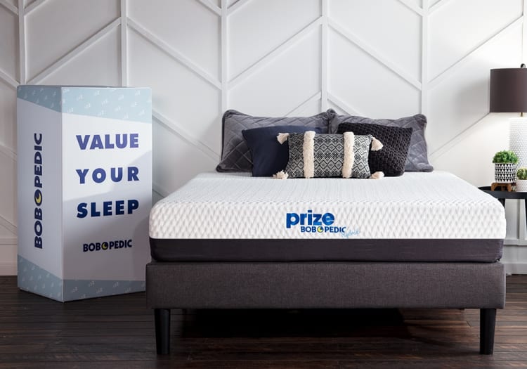 Bob-O-Pedic mattress with white quilted top and dark gray base on a platform bed frame, decorated with patterned pillows. Next to it, a box with text "VALUE YOUR SLEEP" and brand name "BOBOPEDIC" in blue letters.