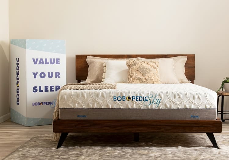 Bob-O-Pedic Sky mattress on a wooden bed frame with beige and white textured pillows, featuring text "VALUE YOUR SLEEP" on packaging box.