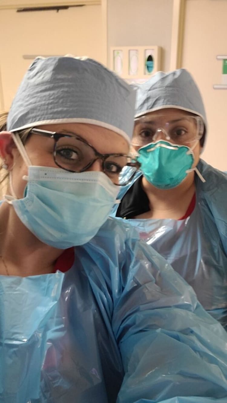 Two healthcare workers wearing light blue disposable surgical gowns, matching caps, and face masks in a clinical setting.