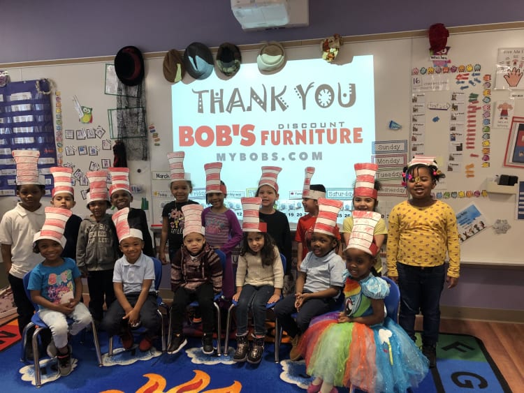 A group of children in a classroom wearing Cat in the Hat striped hats, seated and standing on a colorful carpet. A projector screen behind them displays "THANK YOU BOB'S DISCOUNT FURNITURE MYBOBS.COM" in bold letters. Classroom walls are decorated with educational materials, hats, and charts.
