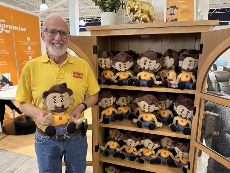 Man wearing a yellow Broy's Furniture polo holding a plush doll with brown hair, yellow shirt, and blue pants, standing next to a wooden cabinet filled with identical plush dolls.