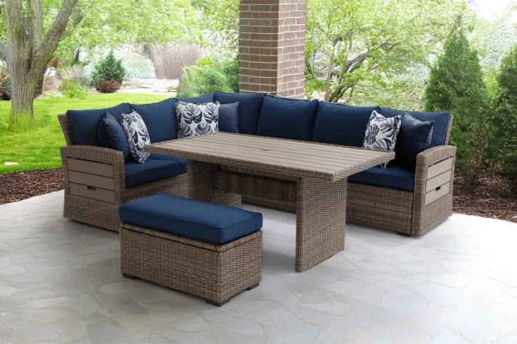 Outdoor patio set with woven rattan sectional sofa, navy blue cushions, patterned throw pillows, matching ottoman, and rectangular wood-look table.