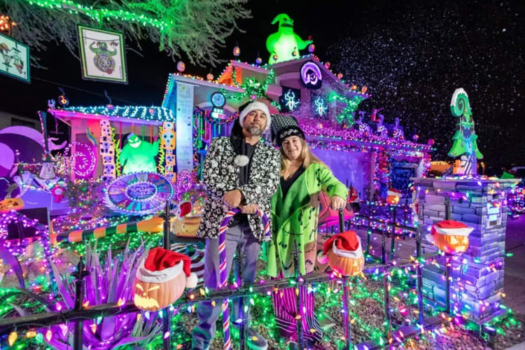 Two people standing in front of a house decorated with vibrant Halloween and Christmas lights, featuring glowing pumpkins with Santa hats, colorful string lights, and festive holiday decorations.