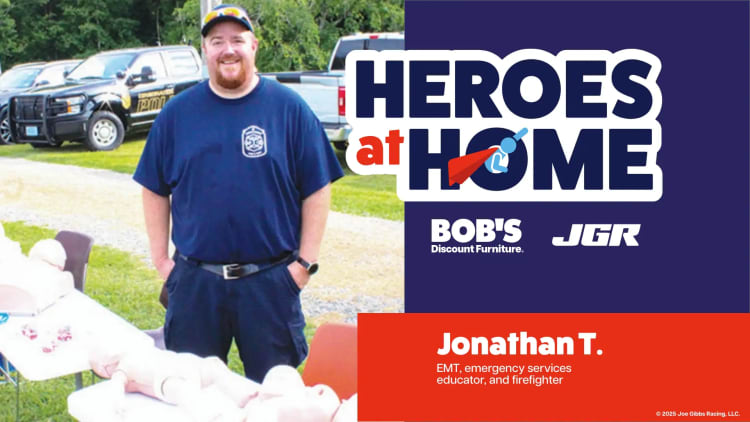 Man in navy blue firefighter uniform stands outdoors next to CPR training mannequins. Text reads: "HEROES at HOME, BOB'S Discount Furniture, JGR, Jonathan T. EMT, emergency services educator, and firefighter. © 2025 Joe Gibbs Racing, LLC."