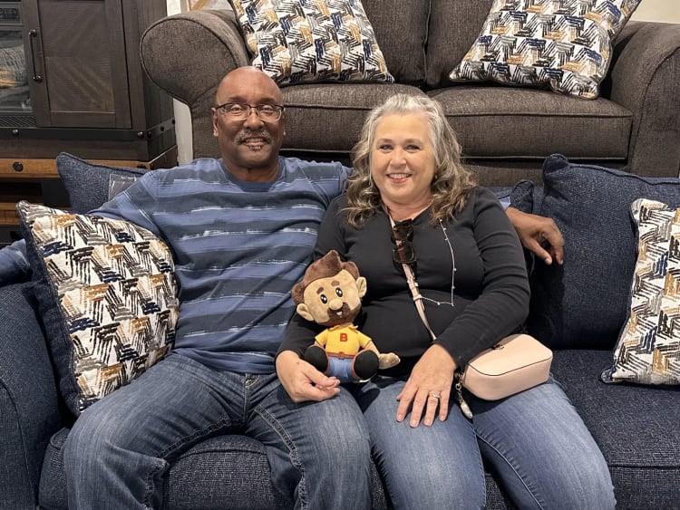 Two people sitting on a blue fabric couch with patterned pillows, holding a plush doll wearing a yellow shirt with a red "B" and blue pants.