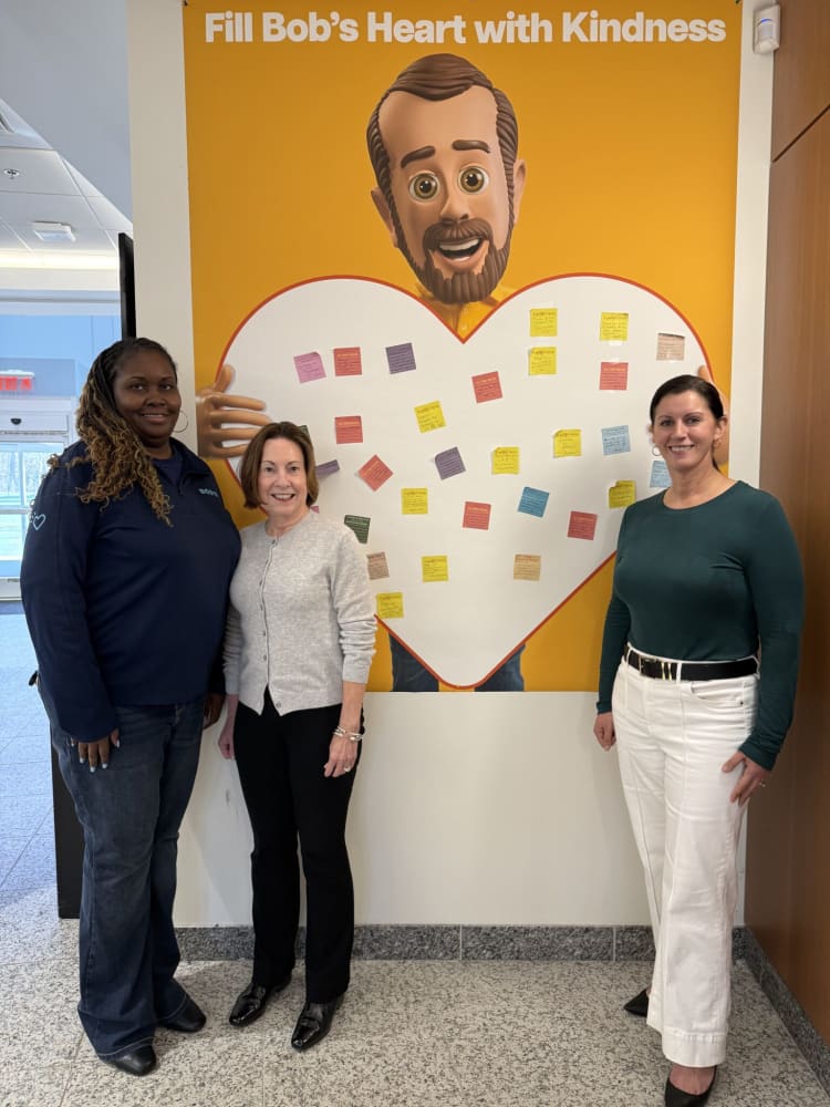 Three women standing in front of a yellow wall display with a large heart and the text "Fill Bob’s Heart with Kindness," featuring colorful sticky notes inside the heart.