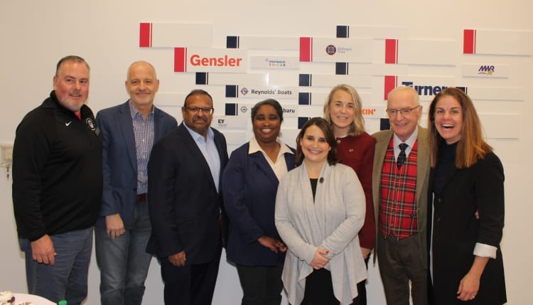 Group of eight professionals standing in front of a wall with logos including Gensler, PepsiCo, Mohegan Tribe, Reynolds' Boats, Subaru, EY, Dunkin', Turner, and MWR, indicating a corporate or networking event.