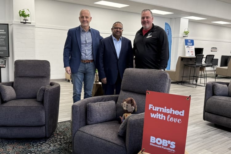 Three men standing behind dark gray upholstered armchairs in a modern furniture showroom. One chair features a small plush monkey and a red sign reading "Furnished with love BOB'S Discount Furniture." The space has light wood flooring, white walls, and office seating in the background.