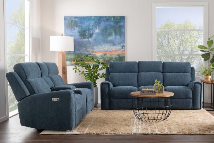 Modern living room setup with blue fabric reclining sofas featuring padded armrests and cup holders, paired with a round wooden coffee table on a beige rug.