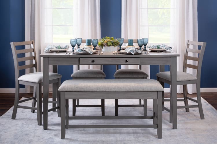 Gray wooden dining table set with cushioned bench and chairs, blue glassware, patterned napkins, and floral centerpiece on light rug.