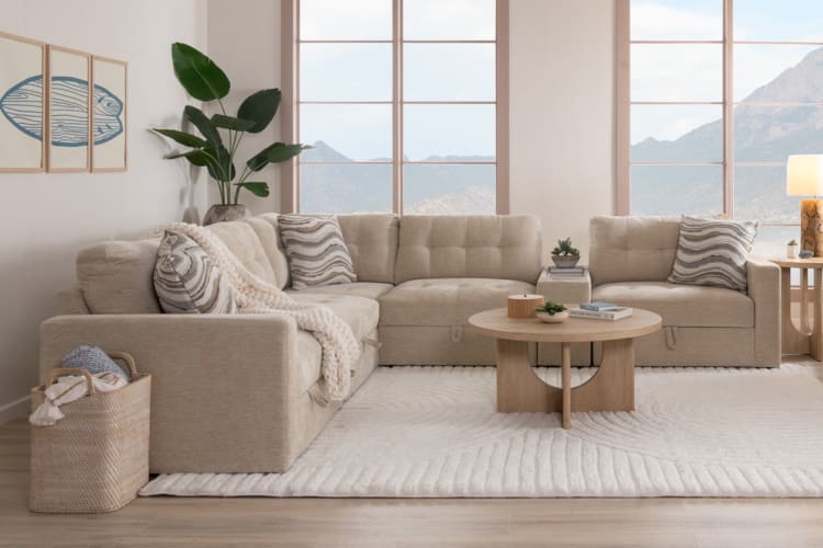 Beige sectional sofa with tufted cushions and patterned throw pillows, paired with a round wooden coffee table on a plush white rug in a bright living room.