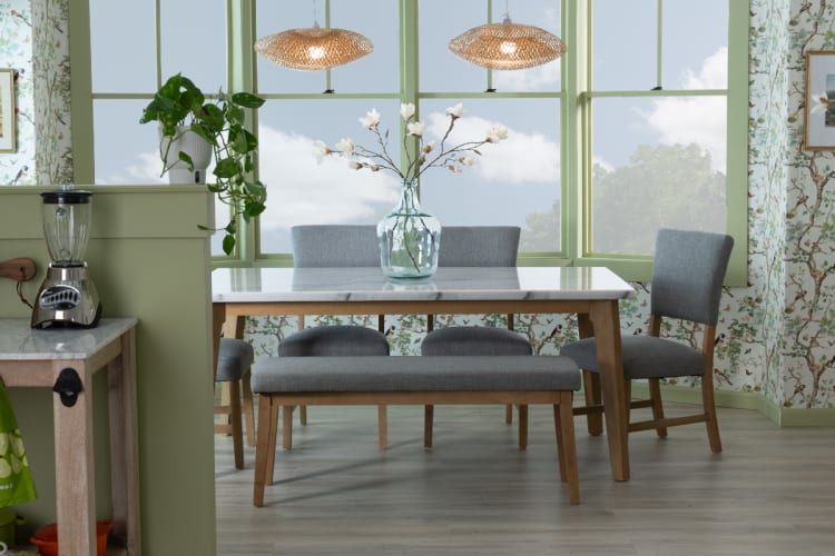 Dining area with a rectangular wooden table featuring a white marble top, gray upholstered chairs and bench, green window frames, floral wallpaper, and hanging wicker pendant lights.