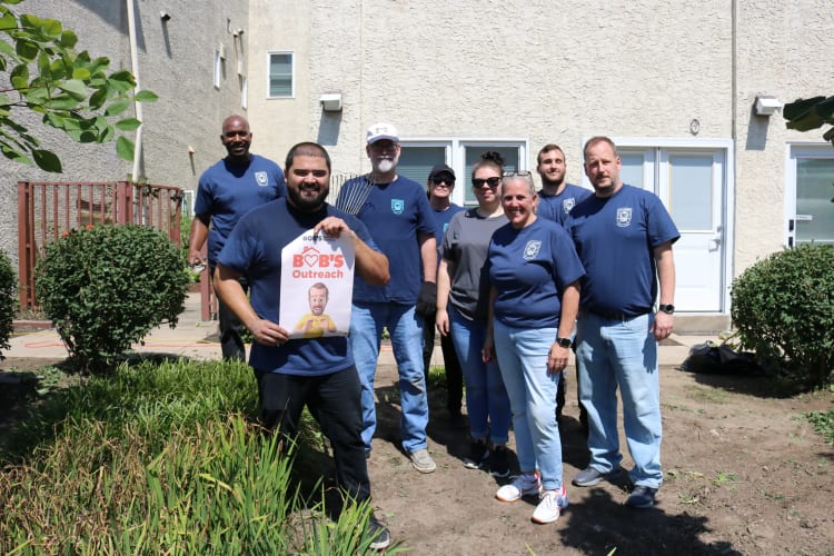 A group of eight people standing outdoors in front of a beige building, wearing blue shirts with a logo, one holding a sign reading "BOB'S Outreach."