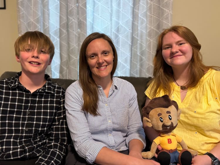 Three people sitting on a couch in a living room, one holding a plush doll with brown hair, beard, yellow shirt with red "B," and blue pants.