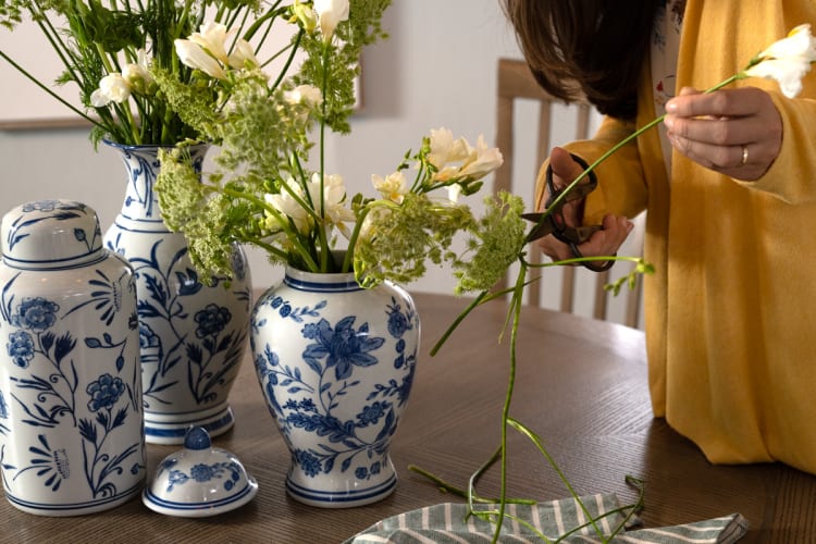 A person in a yellow sweater arranging white and green flowers with scissors in blue and white floral ceramic vases on a wooden table.