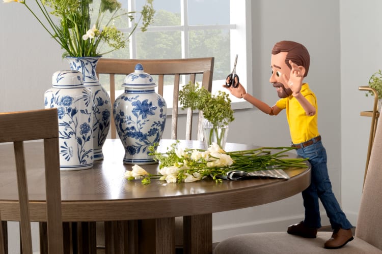 A wooden dining table with blue and white ceramic vases and a bouquet of white flowers, alongside a wooden mannequin holding scissors trimming green stems.
