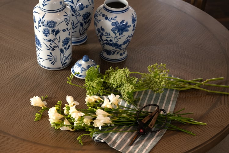 Blue and white ceramic vases with floral patterns on a wooden table, accompanied by fresh white freesia flowers, green foliage, and garden scissors on a striped cloth.