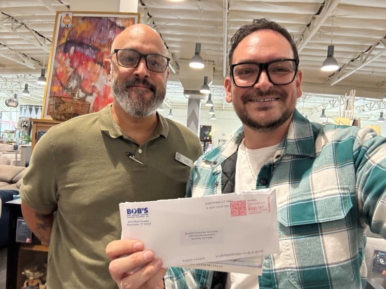 Two men in a furniture store, one wearing an olive green polo with a name tag, the other in a teal and white plaid shirt holding an envelope from Bob's Discount Furniture addressed to Burbank Temporary Act Center, showing a $0.74 postage stamp.