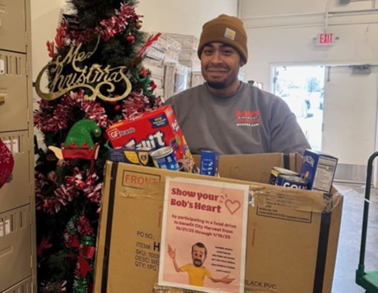 Person holding a box filled with groceries including Fruit Loops cereal, Great Value canned goods, and other food items, standing next to a decorated Christmas tree with a "Merry Christmas" sign. The box has a flyer that reads: "Show your Bob's Heart by participating in a food drive to benefit City Harvest from 10/12/25 through 1/16/26."