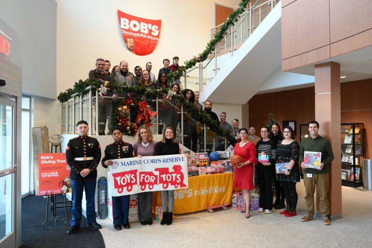 Group of people, including two Marines in uniform, holding a "Marine Corps Reserve Toys for Tots" banner in front of a table with toys and a yellow Bob's Discount Furniture tablecloth, promoting a holiday toy drive inside a decorated building with a staircase and Christmas garlands.
