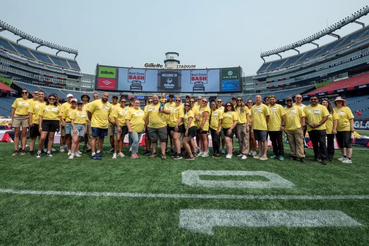 A group of volunteers in yellow shirts standing on the field at Gillette Stadium for the Bed-Zone Bash event, with stadium seating and event signage visible.