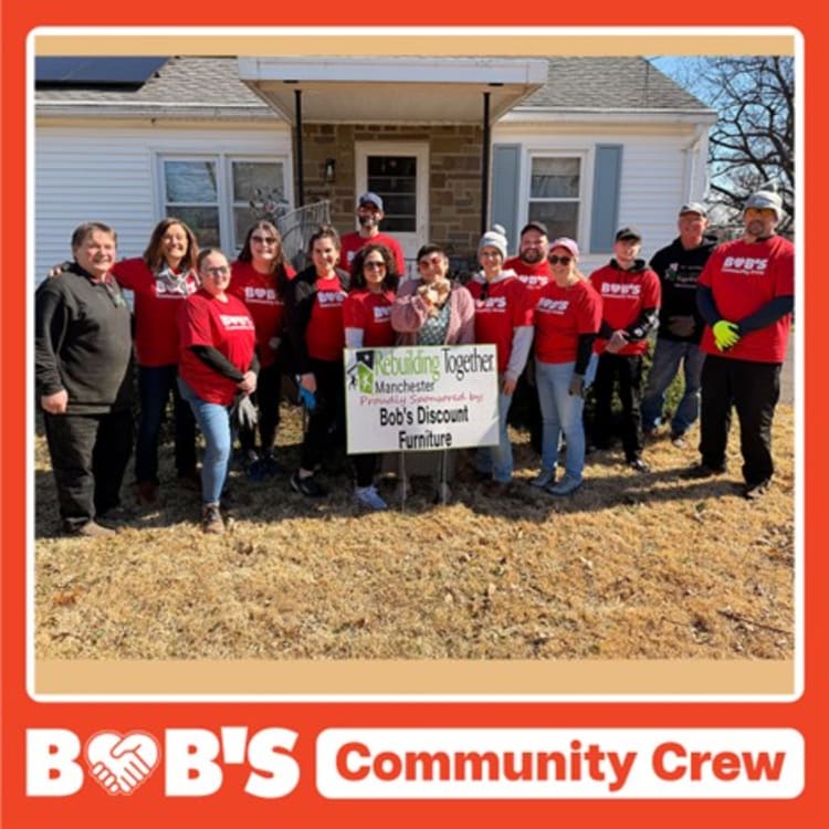 Group of volunteers wearing red Bob’s Discount Furniture shirts standing outside a house holding a sign that reads “Rebuilding Together Manchester Proudly Sponsored by Bob’s Discount Furniture,” promoting community support and volunteerism.