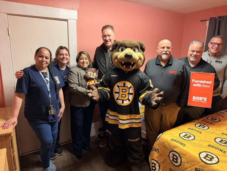 Group of people standing in a room with a Boston Bruins mascot in hockey jersey, a bed with Boston Bruins bedding, and a sign reading "Furnished with love BOB'S Discount Furniture."