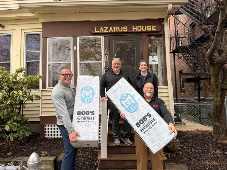 Four people holding large white boxes with blue graphics and text "BOB'S DISCOUNT FURNITURE MYBOBS.COM" stand in front of a building with a sign "LAZARUS HOUSE" above the door, indicating a furniture donation or delivery.