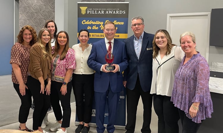 Group of nine people standing in front of a Home Furnishings Association Pillar Awards banner that reads "Celebrating Excellence in the Industry" and "2023 Retailers of the Year," with one person holding a red and clear glass award trophy.
