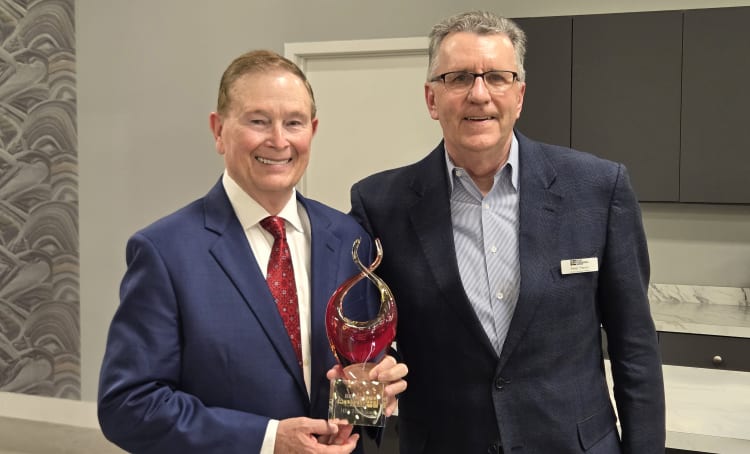 Two men in business suits holding a red and gold glass award trophy with a flame-like design, standing indoors near a marble countertop.