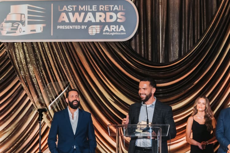 Last Mile Retail Awards stage with three people standing at a podium, backdrop with gold drapes, and a sign reading "LAST MILE RETAIL AWARDS PRESENTED BY ARIA AriaLogistics.com."