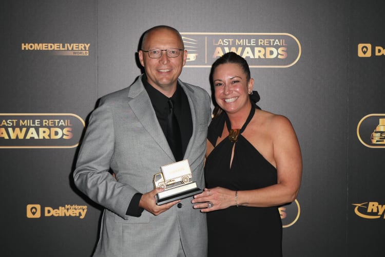 Two people dressed formally at the Last Mile Retail Awards event, holding a gold and black truck-shaped trophy with "AWARDS" text, against a branded backdrop including "HOMEDELIVERY WORLD" and "MyHome Delivery" logos.