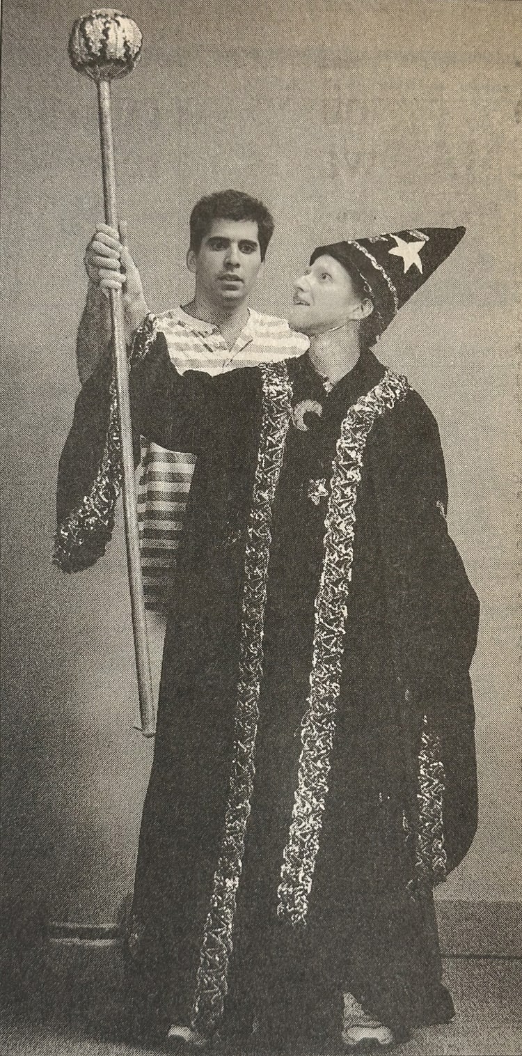 Two people dressed in wizard costumes, one holding a tall staff with a round ornament, wearing a pointed hat decorated with stars and moons, and a long robe with intricate patterns. The other person is standing behind, wearing a striped shirt. The scene suggests a fantasy or magical theme, suitable for costume events or theatrical performances.