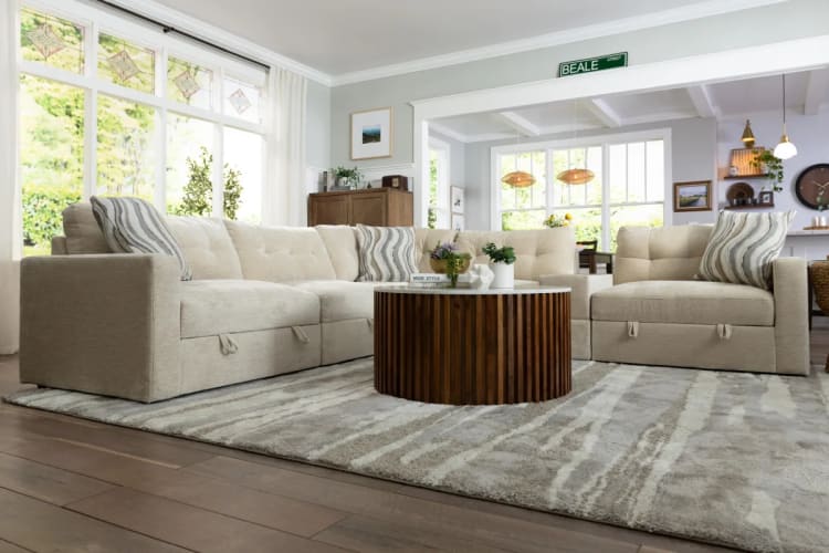 Beige sectional sofa with tufted cushions and striped throw pillows arranged in an L-shape on a patterned gray rug, centered around a round wooden coffee table with white marble top, set in a bright living room with large windows and light gray walls.