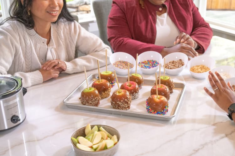 Caramel apples on a baking tray with colorful sprinkles and chopped nuts, surrounded by bowls of toppings and sliced apples on a marble table.