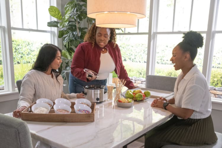Three women gathered around a marble kitchen table with a slow cooker, bowls of colorful sprinkles, and fresh fruit, preparing a dessert.