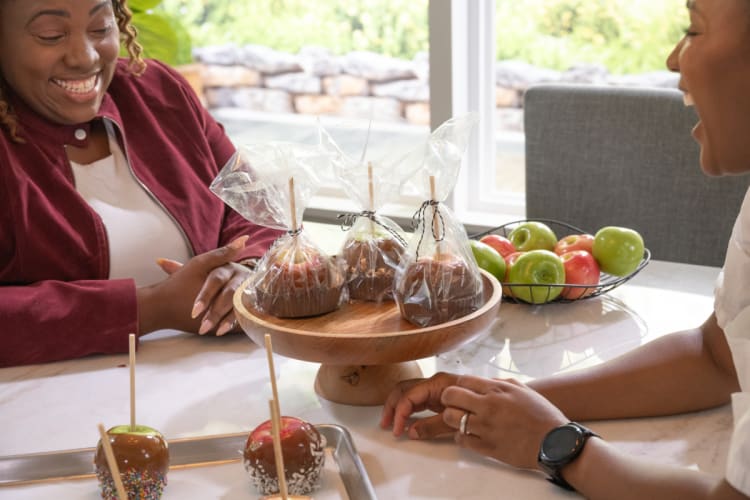 Two people smiling and sitting at a table with caramel apples and chocolate-covered apples on wooden and metal trays, with a bowl of red and green apples in the background.