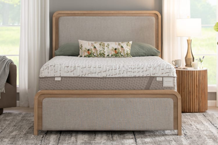 Modern bed with light wood frame and beige upholstered headboard and footboard, featuring a plush mattress labeled "Enchant" with a white textured cover and floral decorative pillow, set in a bright bedroom with natural light and wooden nightstand with lamp.